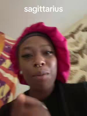 A close-up shot of a person with dark skin, viewed from the chest up. The person is facing the camera, and their facial expression suggests a sense of contemplation or mild humor. They are wearing a dark-colored top and a bright pink head covering. The background shows a mix of blurred colors which might suggest a patterned fabric or wall decor.