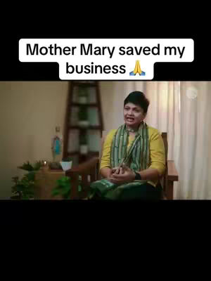 The cover image shows a woman sitting indoors, likely in a home or studio setting. She is the central subject, positioned in front of a neutral background with soft lighting and a serene atmosphere. She is sitting up straight, engaged in conversation, and a text overlay stating 'Mother Mary saved my business' is above her, communicating the video's core message. Overall, the composition is simple, with the woman and the text overlay as the main focus.