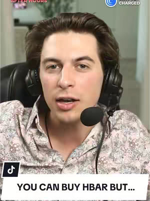 A man with brown hair and a headset is centered in the frame. He is wearing a patterned button-down shirt and looking directly at the camera. A microphone is positioned near to his mouth. The background is blurred, but a glimpse of an interior setting is visible.