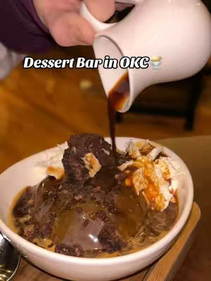 A close-up shot captures a bowl of dessert being drizzled with sauce from a white pitcher held by a person, with a dark and luscious dessert. The bowl is white, and the lighting is warm.