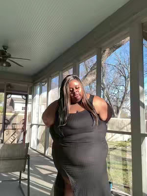 A woman stands in a sunlit room with large windows and a view of trees, wearing a black dress. The lighting suggests early morning or late afternoon, creating soft shadows. The background includes a ceiling fan and what seems to be an outdoor porch. The composition is straightforward, with the subject centered.