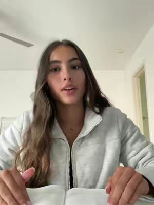 A young woman with long brown hair is centered in the frame, giving a monologue while looking into the camera. She wears a light grey zip-up jacket with a black zipper. The background is a light-colored wall, ceiling, and door.