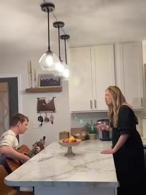 A woman with long blonde hair wearing a yellow headband and a black dress is singing standing next to a kitchen island. Behind her is a man who appears to be concentrating while playing an acoustic guitar. The kitchen is bright and has white cabinets. The lighting is provided by hanging lights and it is a medium shot.