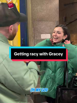 A candid shot captures a man and a woman in an outdoor setting. The man, seen from the side, is wearing a dark hat, and an olive green jacket, with a vibrant yellow sign peaking out from behind his right shoulder. The woman, angled slightly, is wearing a green puffer jacket and appears to be reacting enthusiastically, her face lit with a joyful expression. The background consists of a building facade.