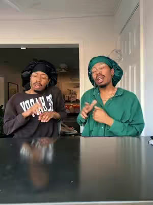 Two people, both wearing matching head coverings and casual attire, are positioned to the left and right of a dark surface. The left individual is wearing a black sweater with white block letters; the right is in a green shirt. Both are making hand gestures and appear to be in conversation. The background includes a bright opening to another room and a plain, off-white door.