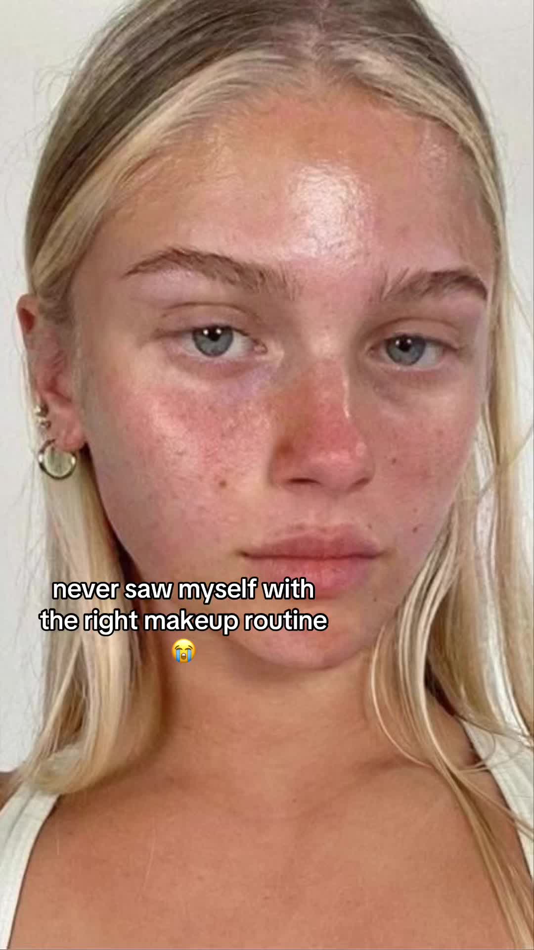 A close-up shot featuring a young woman with fair skin, light blonde hair, and blue eyes. The image is brightly lit, emphasizing the details of her face, with some redness on her cheeks and nose. Written text appears at the bottom describing the makeup routine.
