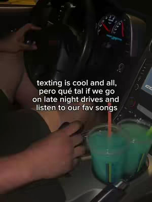 An overhead shot inside a car. The steering wheel and dashboard dominate the scene, the creator’s hands visible holding the wheel and a drink. Text overlayed: 'texting is cool and all, pero qué tal if we go on late night drives and listen to our fav songs'. The car's interior is dark, contrasting with the bright drinks in the cup holder.