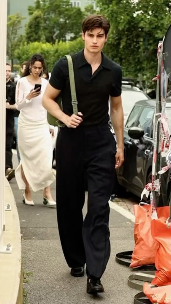 A well-dressed man walks on a sidewalk near parked cars. He wears a black polo shirt, dark trousers, and carries a green bag. In the background, a woman in a white dress and a few other people are visible on a street.