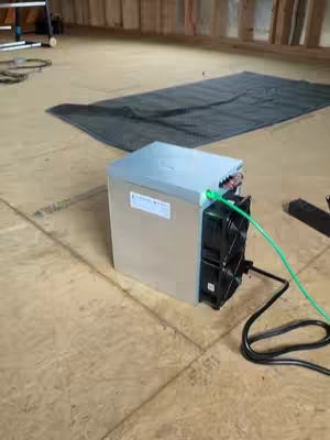 The image is a medium shot of a Bitcoin mining device set up in an unfinished looking room with wood paneling and a black mat on the floor. The device is a metallic silver squared box with a black fan visible on the side facing the camera. It is connected to a green and black cable. The overall tone is practical and informational, likely showcasing cryptocurrency mining.