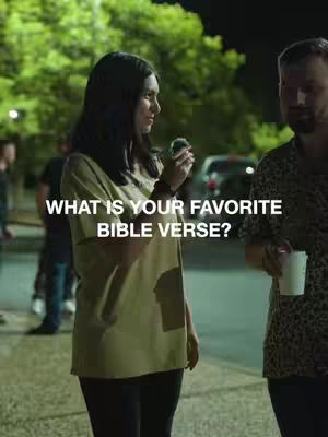 A woman in a casual green shirt holds a small object, seemingly talking with another person, with a blurred background featuring more people and foliage, at dusk or nighttime. Text overlayed reads, 'What is your favorite Bible verse?'