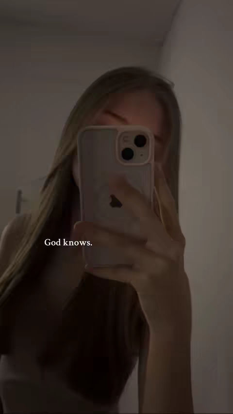 A woman is taking a selfie in front of a mirror. She's holding up a phone that is covering most of her face. The background is plain wall. The image has soft lighting.