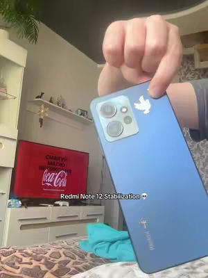 A person holds a blue smartphone with a camera module, with a white dove sticker on the back. The background shows a living room scene with a TV displaying a Coca-Cola ad. The foreground has white furniture and part of a bed. The video title appears below the phone.