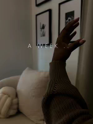 The image shows a person's hand reaching up, with a soft background of a neutral-toned room. The hand is in the foreground, its fingers slightly curved. The background includes a white pillow, a neutral-colored sofa, framed pictures, and a wall. The overall lighting is soft and natural. A text overlay across the center reads "A WEEK ..".