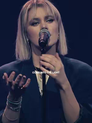The cover image features a person with blonde hair and a microphone, seemingly singing or performing on stage. The person is centered, with their hands raised and open, and a text overlay that reads 'COME INTO THE LIGHT.' The background is dark, and the lighting suggests a stage or studio setting.