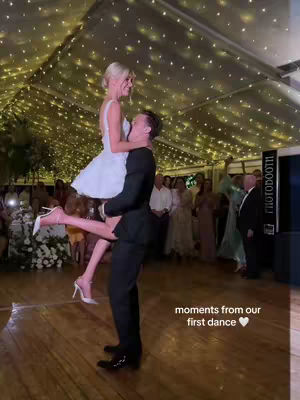 A couple is shown during their wedding reception's first dance. The bride is clad in a short white dress and is lifted by the groom, who is wearing a black suit. They're on a wooden dance floor under a tent decorated with string lights. Attendees watch in the dim lighting.