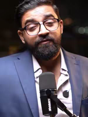 The cover image shows a man in a blue blazer and a white shirt, wearing glasses and holding a microphone. He has a beard and appears to be speaking. The background is dark, and the lighting appears to be artificial.