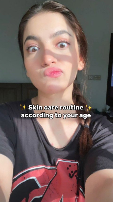 A young woman with light skin and dark hair is in a selfie-style video. She has her hair pulled back in a braid. She has a surprised expression, with her eyes wide and her mouth pursed as if she is blowing a kiss. She is wearing a black t-shirt with a red design on it.
