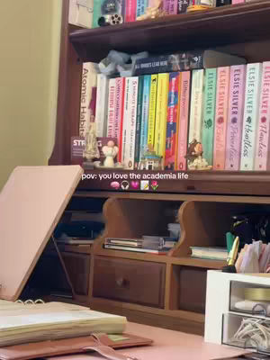 A well-lit, cozy room interior featuring a desk with an open book and a laptop, a bookshelf filled with books and decorative items, and a small statue. The scene is dominated by warm, inviting tones and a focus on study or work.