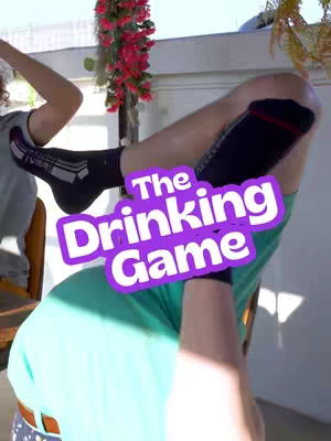 A person lying on the floor with their legs in the air, over them is a purple and white graphic that says 'The Drinking Game'. The person is wearing a green t-shirt, shorts, and black socks.