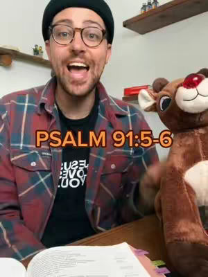 A man wearing glasses and a beanie smiles directly at the camera with an open mouth. Behind him is a stuffed reindeer and on the image overlayed is the text "Psalm 91:5-6". He is sitting at a table with a book open in front of him.