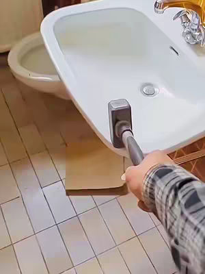 The image shows a hand holding a cleaning tool to scrub a white ceramic sink. The hand is wearing a plaid shirt. The background includes a white toilet and gray tiled flooring.