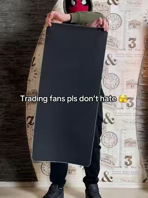 A person, dressed in a black outfit and a hood, is holding a large, black rectangular object in front of them. They are wearing a mask with a red face and a black spiderweb design. The background has patterned wallpaper with clocks, and the text 'Trading fans pls don't hate' is overlaid on the image.