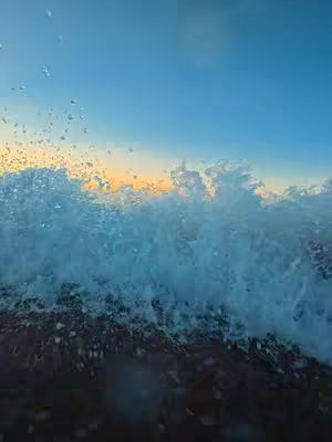 The cover image depicts a dynamic seascape. A large wave is crashing, with the foamy white crest of the wave dominating the center of the frame. Water droplets are suspended in the air, captured at the peak of the wave's action. The light from the setting sun is illuminating the wave crest, creating a gradient that moves from a rich blue in the sky to an orange-yellow towards the horizon. The color palette consists primarily of blues, whites, and the warm tones of the sunset.