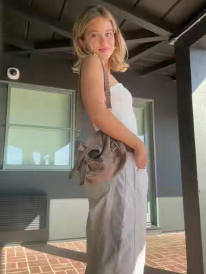 A young woman stands outdoors, angled slightly towards the camera, wearing a white top and light-colored pants, with a cross-body bag. She has blonde hair and a subtle smile. The background shows a dark exterior wall with windows and a brick patio.