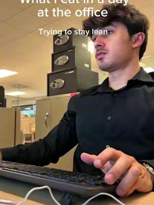 A man sits at a desk in an office setting, a keyboard in front of him. Above his head and to his left are stacks of black file cabinets. He is wearing a black shirt.