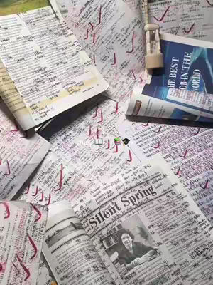 A cluttered workspace with open books and papers spread across a surface. The papers are covered in handwritten notes, annotations, and red check marks, suggesting a study environment. A small hourglass is placed amongst the papers. The overall impression is of intense study or review.