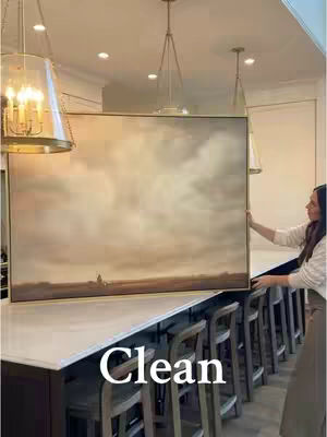 A woman standing in a modern kitchen holding a large, framed painting. The kitchen features a long island with stools, overhead lighting, and a neutral color palette. The painting depicts a landscape with soft, cloudy skies.