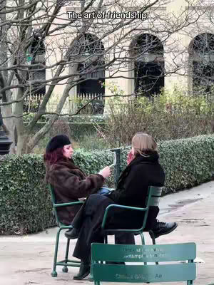 Two women are seated in a park, facing each other. The woman on the left is wearing a black beret and brown fur coat, and the woman on the right has blonde hair and is wearing a black jacket. They are seated on chairs with a green structure, set against a background of trees and a building with arched windows.