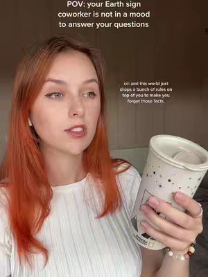 A woman with red hair and fair skin is centered in the frame, holding a beverage container. Text overlays are present, and the background appears to be a light-colored wall. The woman is smiling and looking towards the camera.