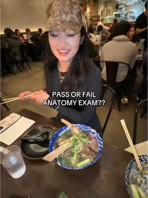 A woman in a camo hat smiles while holding chopsticks at a table in a restaurant. A bowl of food is in front of her, and a text overlay asks about passing or failing an anatomy exam. Other people are visible in the background.