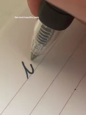 A close-up shot shows a hand holding a clear pen, writing on lined paper. The text overlay reads 'the most beautiful pens'. Dark ink forms the number '2' on the page. The background is slightly blurred, suggesting a casual setting.