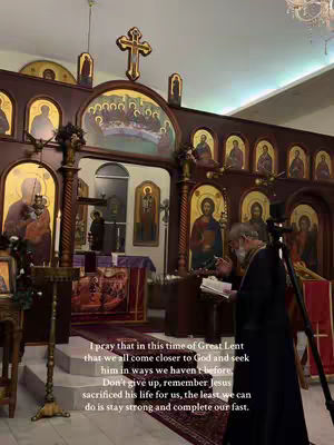 An interior shot of a church during a religious service. A man in a dark robe is reading from a book, a decorated altar is in the background, and religious icons and artwork adorn the walls. The lighting is dim, suggesting a solemn mood.