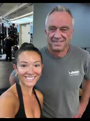 A selfie-style shot taken inside a gym. The primary focus is on a woman in a black workout top and a man in a gray t-shirt, both smiling broadly at the camera. The woman, positioned in the foreground, features in-focus, with a clean, slightly out-of-focus gym environment in the background with exercise equipment.