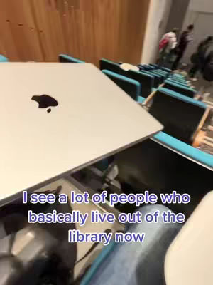 A first-person perspective shot inside a library, showcasing rows of study carrels, a laptop with the Apple logo open on the desk, and blurred figures in the background. Text overlayed on the image reads 'I see a lot of people who basically live out of the library now' in white font.