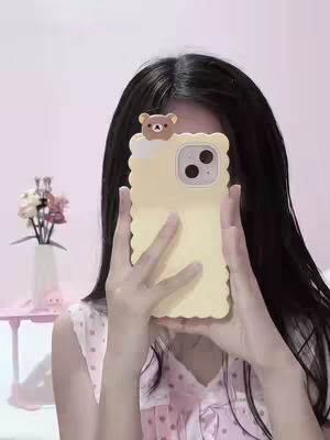 A person, likely a young woman, is holding a light yellow phone case up in front of their face, obscuring most of it. Her dark hair is long and falls down both sides of her face. The phone case is decorated with a small Rilakkuma bear figure at the top. The background appears to be a soft light pink, with a glimpse of a table holding a vase of flowers on the left side.