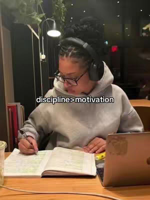 A young woman with dark hair, wearing glasses and headphones, sits at a table in front of an open notebook and a laptop. She is writing with a pen, with a cafe-like setting in the background.