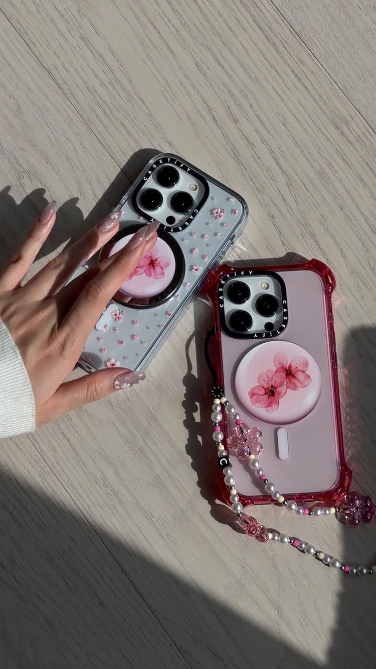 A close-up shot showcases two iPhones with stylish cases featuring floral designs. One phone is on a wooden surface. A hand with manicured nails is positioned near a phone. The lighting is bright, casting shadows that enhance the visual elements.