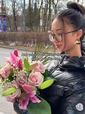 A young woman in a black puffer jacket holds a large bouquet of flowers in front of a blurred outdoor background. The flowers include pink roses, lilies, and other greenery. The woman wears glasses and has her hair up. Text overlays the image.