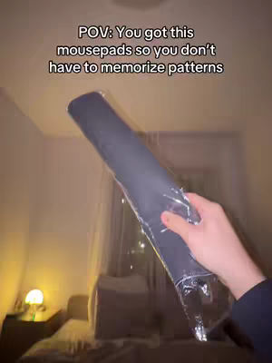 The image shows a person's hand holding a rolled-up, long object, possibly a mousepad, inside a transparent plastic wrapper. The background is a dimly lit room with items like a table lamp and bed, appearing to be a bedroom setting. The lighting is warm, and the overall composition focuses on the object being held.