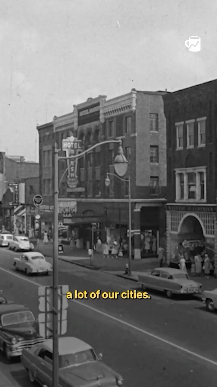 A black and white photograph captures a street scene from what appears to be the mid-20th century. The perspective is focused on a city street lined with buildings, including a prominent multi-story building with a hotel sign. Classic cars are parked along the street. The sky is overcast, contributing to a vintage and nostalgic feel.