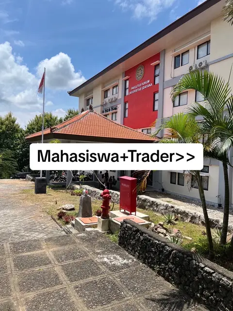 The image shows a university building with a white rectangular text overlay. The building has a red portion, a tile roof, and a flag. There's a clear blue sky with clouds. In the foreground, there are steps, a red fire hydrant, and greenery.