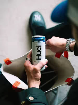 The image shows a person holding a can of 'Huel' energy drink inside an open tote bag. The person is wearing a dark jacket and a silver watch with a silver band. The background is blurred, suggesting an outdoor or public setting.