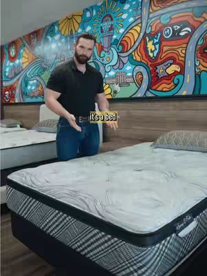 A man stands in front of a bed in a showroom, holding a sign that says 'it's a bed.' The background features a colorful mural. The man wears jeans and a black shirt.