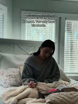 A woman sits on a bed, dressed in a gray sweater and gazing downward, holding a pen. A book is open on the bed, near a pink item, with a window and blinds in the background. The scene is dimly lit.