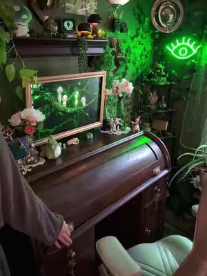 The image shows a cozy, green-themed gaming setup with a roll-top desk. The desk is made of dark wood, and the room is filled with decorative items like plants, artwork, and fairy lights. The overall composition is warm and inviting, with a focus on creating a comfortable and aesthetically pleasing environment.