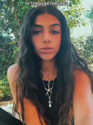 A woman with long, dark wavy hair is centered in the frame. She wears layered necklaces and a black top. The background is blurred, with green foliage visible. Text overlayed at the top reads 'AFTER HEARTBREAKS'.
